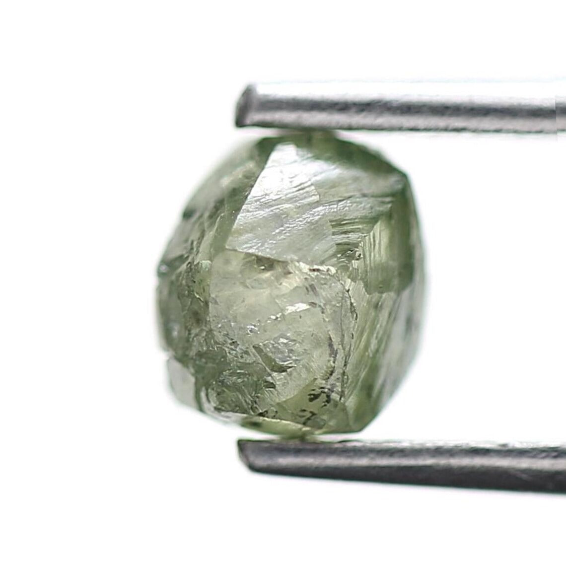 A faceted, rough-cut light green diamond held in silver metal tweezers against a plain white background.